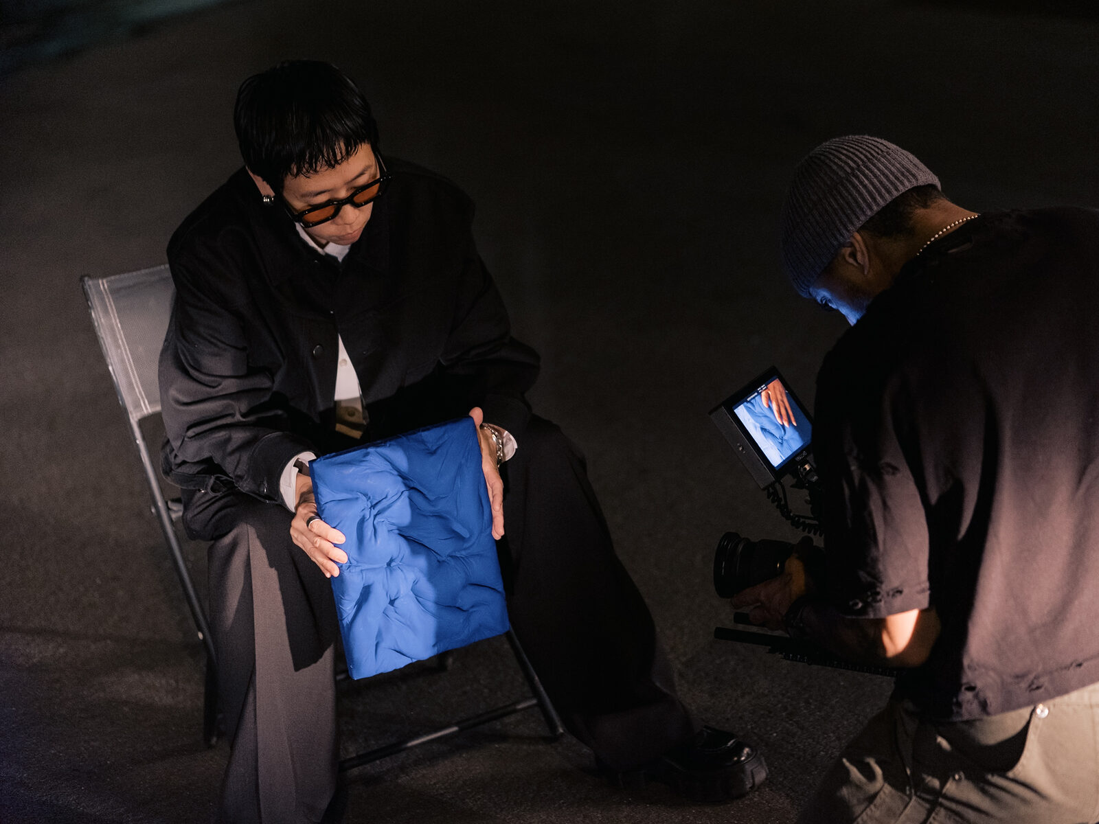 A seated artist holds a blue relief panel while a crew member checks the camera monitor