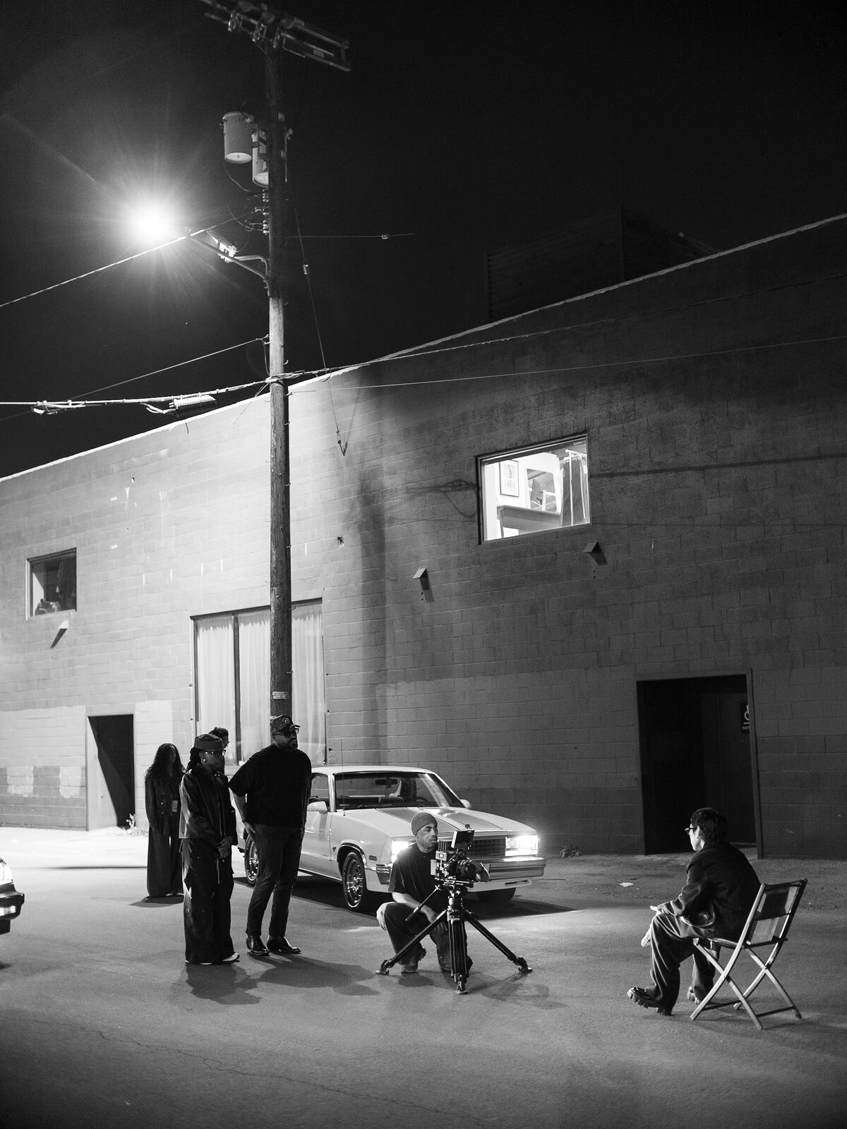 Black and white frame of the crew lighting a seated subject beside a car under a streetlight