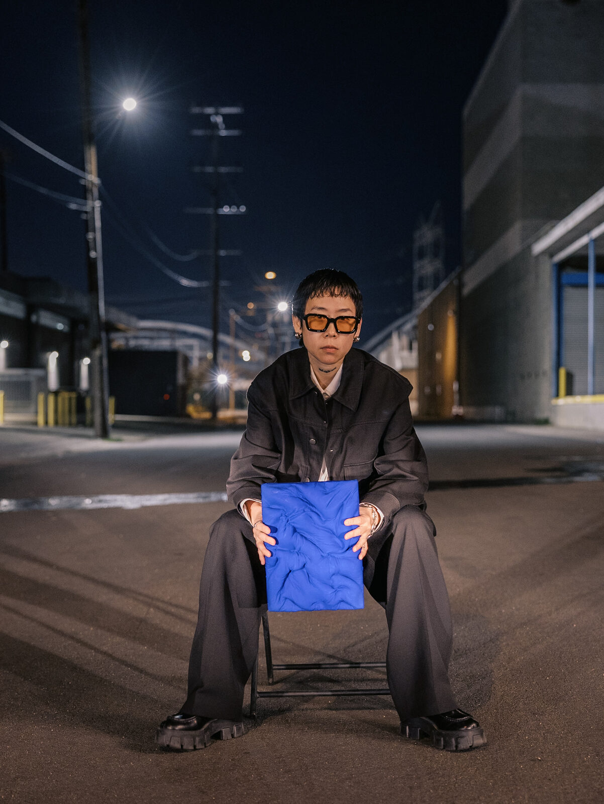 An artist seated on a folding chair holding a blue relief panel between takes